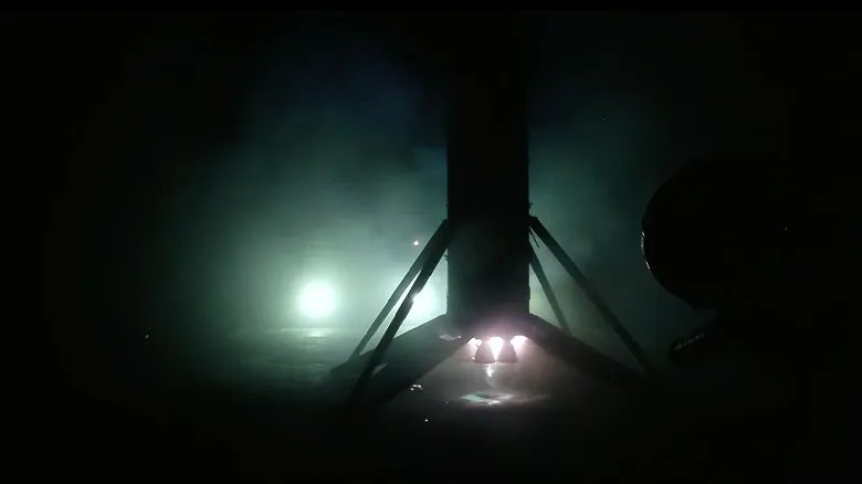 the first stage of an orbital rocket is seen in silhouette backlit by spotlights as it stands upright on its landing legs atop an ocean-based droneship