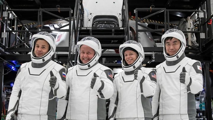 three men and one woman all in white spacesuits give a thumbs-up in a training facility here on earth